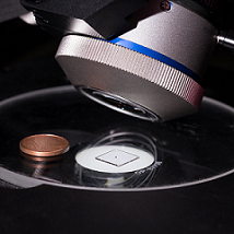 University of Stuttgart researchers used a microscope to acquire tilted-view images of a 600-µm-diameter doublet lens system that was 3D-printed on a 1x1 cm² glass slide. The doublet lens system is visible as the small dot in the center of the glass slide. The coin is included for scale. Courtesy of Moritz Flöss, University of Stuttgart.
