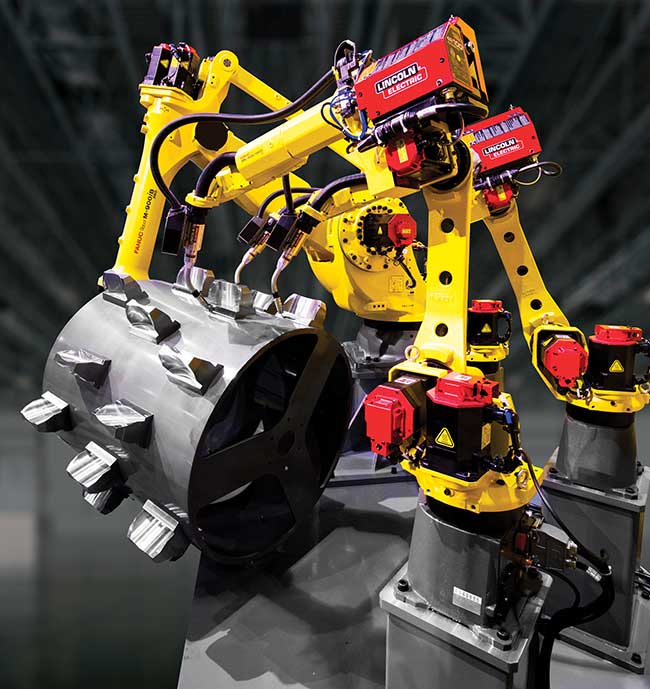 A FANUC M-900iA/360 robot picks up a compactor drum and presents it to three ARC Mate 100iC robots equipped with iRVision to locate parts for welding.