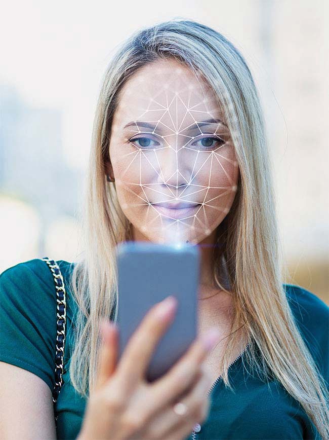 A representation of an NIR system being used for facial recognition. The smartphone emits a diffractive optical element (DOE) dot-grid pattern (invisible to the user) that is cast on the face. An IR camera inside the phone captures the reflection of NIR light and analyzes deformations of the grid pattern to determine 3D contours of the face for identification.