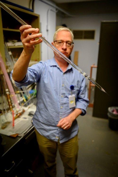 Colin Baker, U.S. Naval Research Laboratory, holds a silica glass rod (optical preform) that will be pulled into an optical fiber suitable for production of an eye-safer laser. Courtesy of Jonathan Steffen/U.S. Naval Research Laboratory.