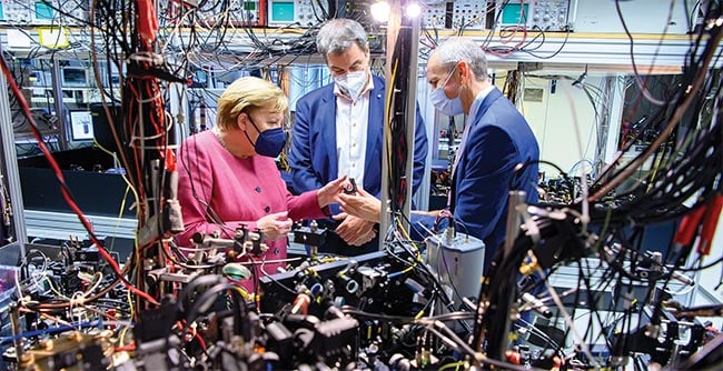 Figure 3. Integration and miniaturization are problematic for quantum computing components that are based on trapped ions and cold atoms, as illustrated by a setup shown to Germany’s then-Chancellor Angela Merkel and Bavarian Minister-President Markus Söder during a September 2021 tour of the Max Planck Institute of Quantum Optics (above). Courtesy of Max Planck Institute of Quantum Optics