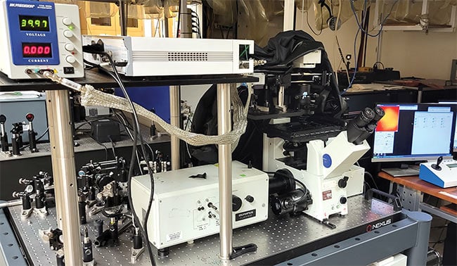 A laser-scanning stimulated Raman spectroscopy (SRS) microscope setup at the University of California, Irvine is powered by a picosecond light source, a lock-in amplifier for isolating the SRS signal, and a computer for collecting imaging data (top). Advanced techniques such as SRS have overcome some of the traditional challenges of spontaneous Raman emission to become new tools with which to monitor cellular processes.  An SRS microscope image of a cultured human lung A549 cell (bottom). The image is segmented into two principal components, which represent the presence of organic matter (blue) and water (green). Courtesy of Courtesy of University of California, San Diego/Judy Kim.