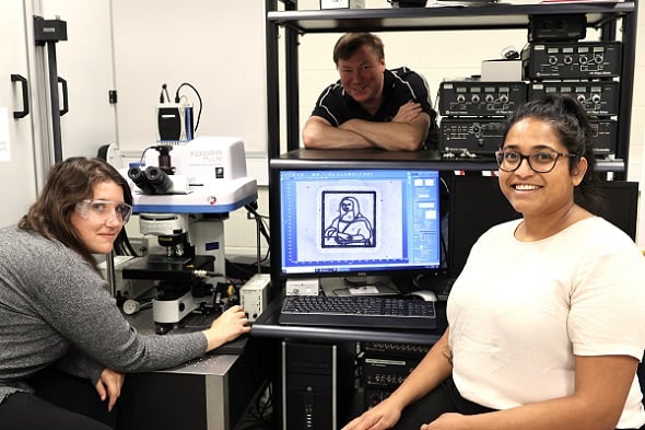 Researcher Abigail Mann (left) next to the low-power laser, Australian National Fabrication Facility spectroscopist Jason Gascooke (center) and researcher Lynn Lisboa (right) with the “micro-Lisa” laser image displayed on a regular computer screen. Courtesy of Flinders University.